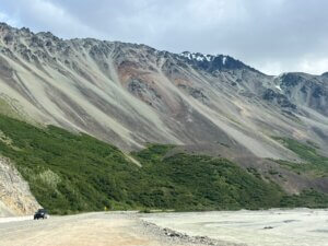 Samochód jadący górską drogą wzdłuż rzeki w surowym, skalistym krajobrazie Alaski z zielonymi zboczami i stromymi szczytami w tle